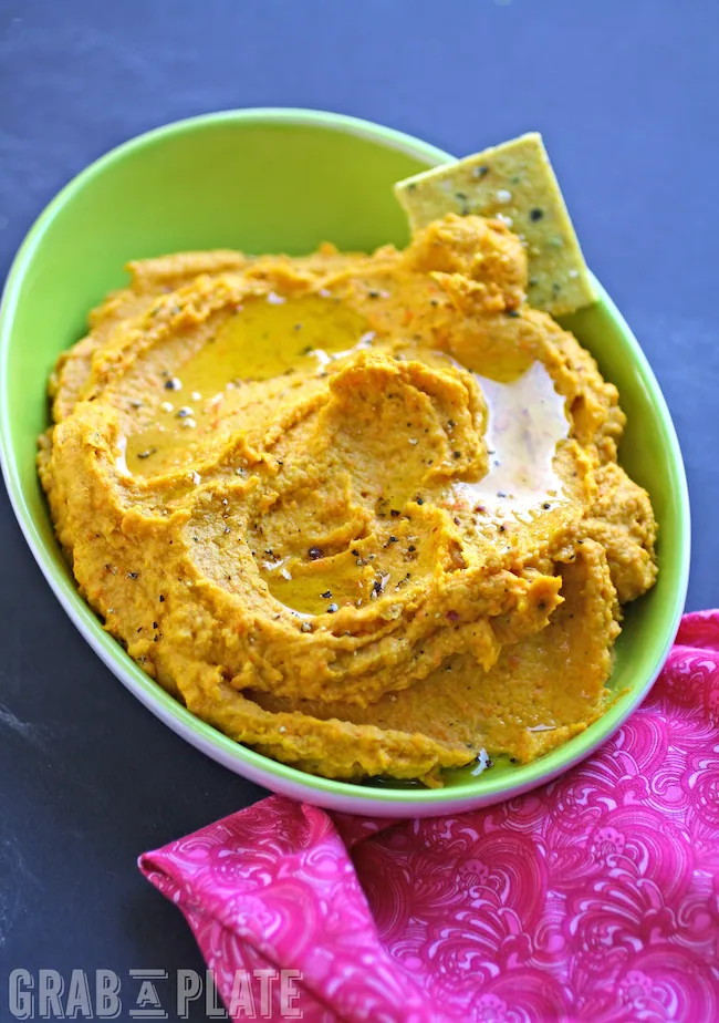 A bowl of colorful Carrot Hummus served with Curried Hemp Seed Crackers