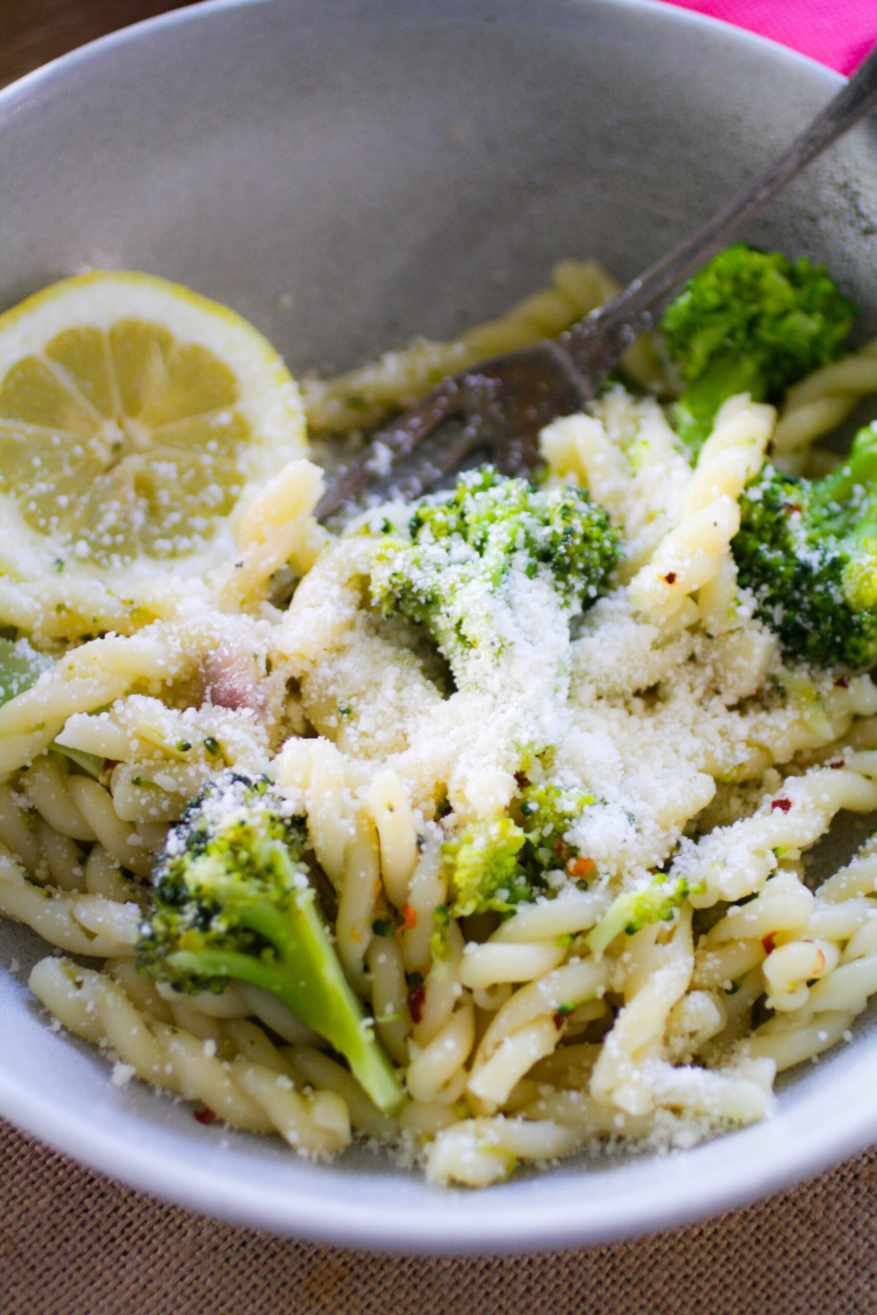 A bowl of spiral pasta with broccoli florets mixed in and covered in Parmesan cheese and a lemon slice on the side.