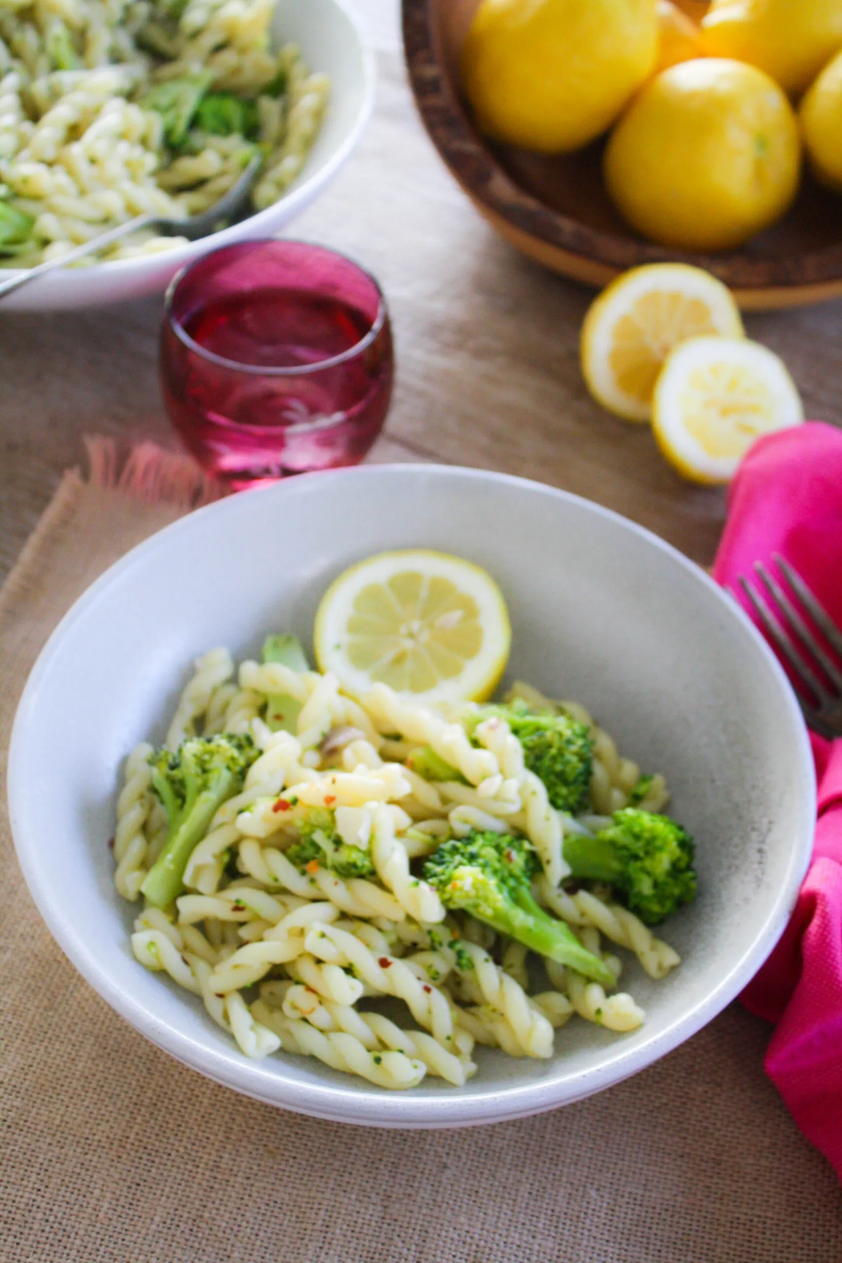 A table with a bowl of spiral pasta and broccoli florets mixed in with a red glass, pink napkin, and lemon slices.
